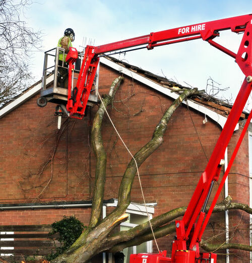 Tree surgeon working up cherry picker repairing storm damaged roof after an uprooted tree fell on top of a residential house